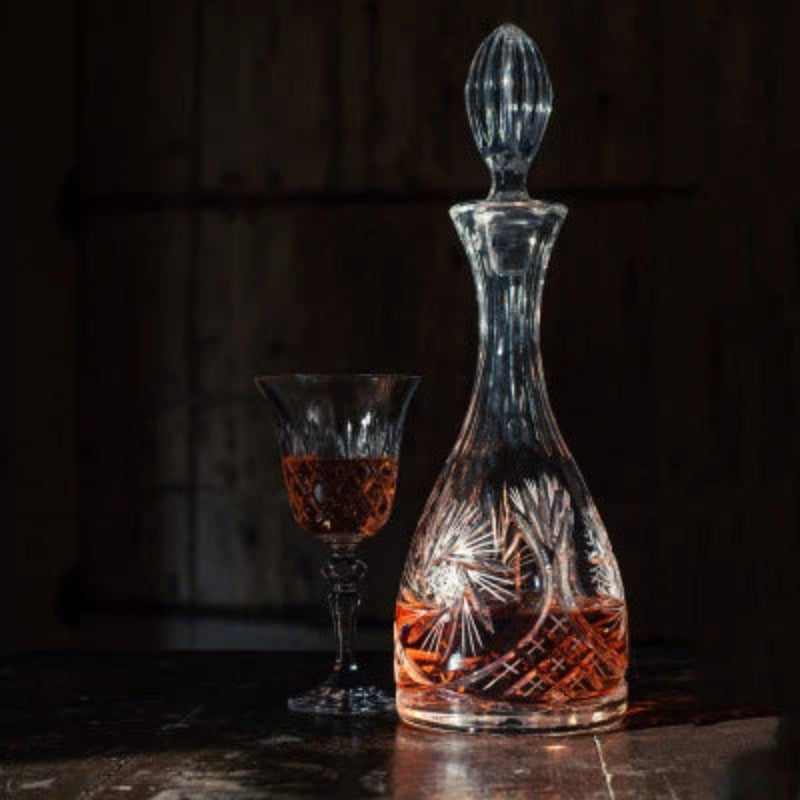 A crystal wine decanter with a teardrop-shaped body featuring intricate cut patterns, topped with an elongated, faceted stopper.