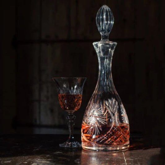 A crystal wine decanter with a teardrop-shaped body featuring intricate cut patterns, topped with an elongated, faceted stopper.